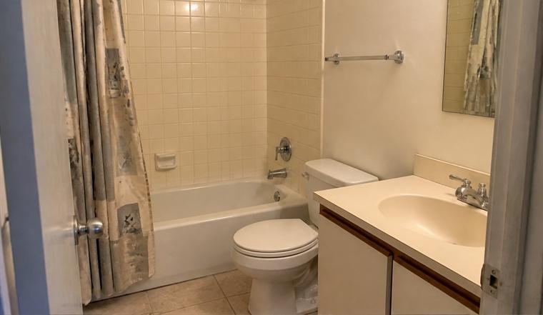 Bathroom with yellowed square tile, a worn shower curtain and a cream laminate vanity, before renovation.